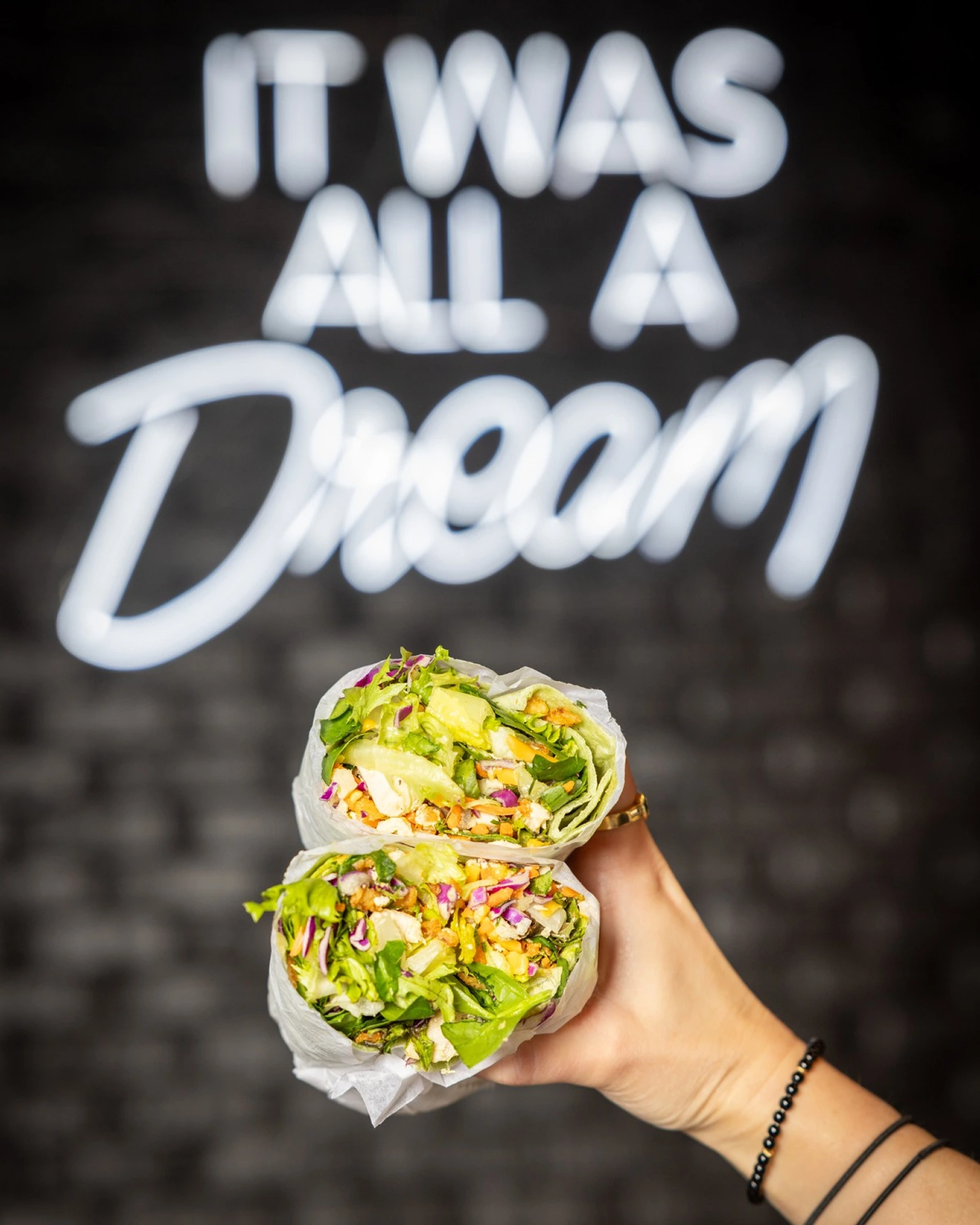 A hand holding a wrap cut in half, in front of a black wall with neon spelling 'It was all a dream'
