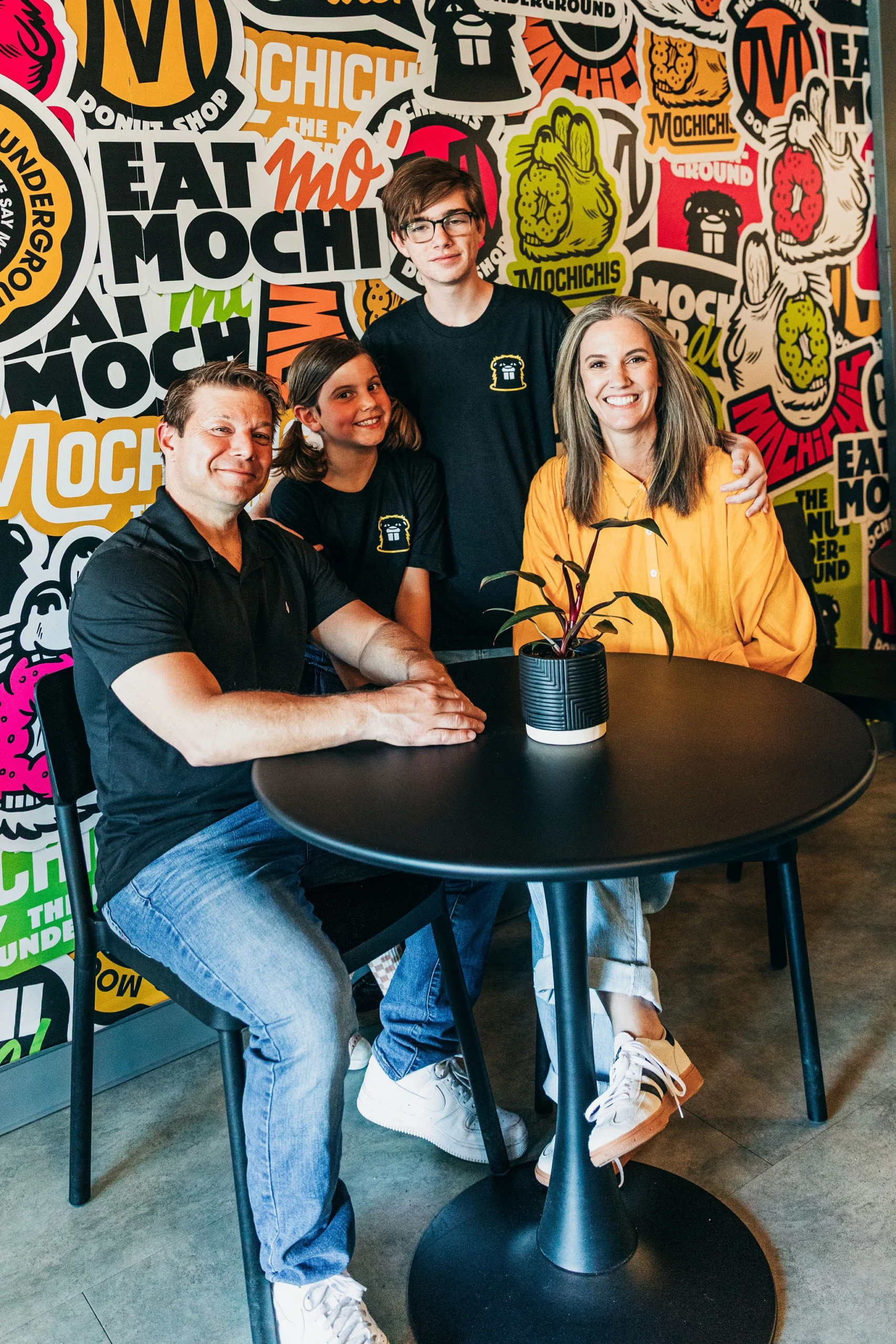 The owners of Mochichis Donut Shop and their family sitting in front of a colorful sticker mural the shop