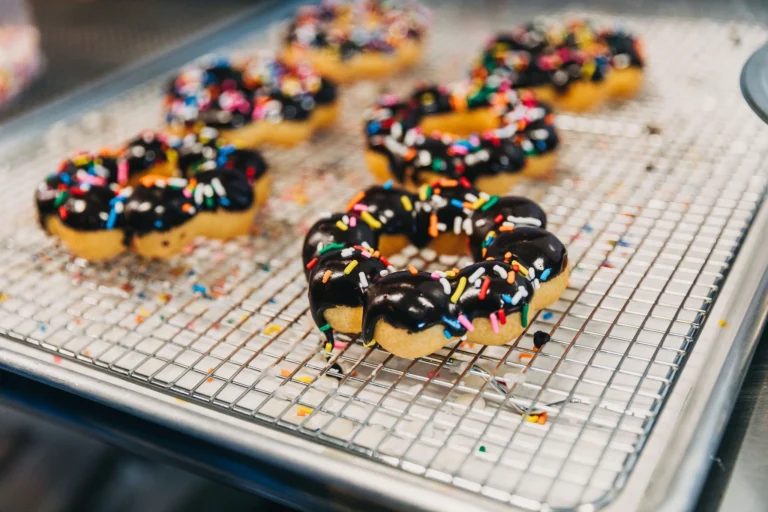 Fresh mochi donuts dipped in chocolate and covered in rainbow sprinkles on a cooling tray