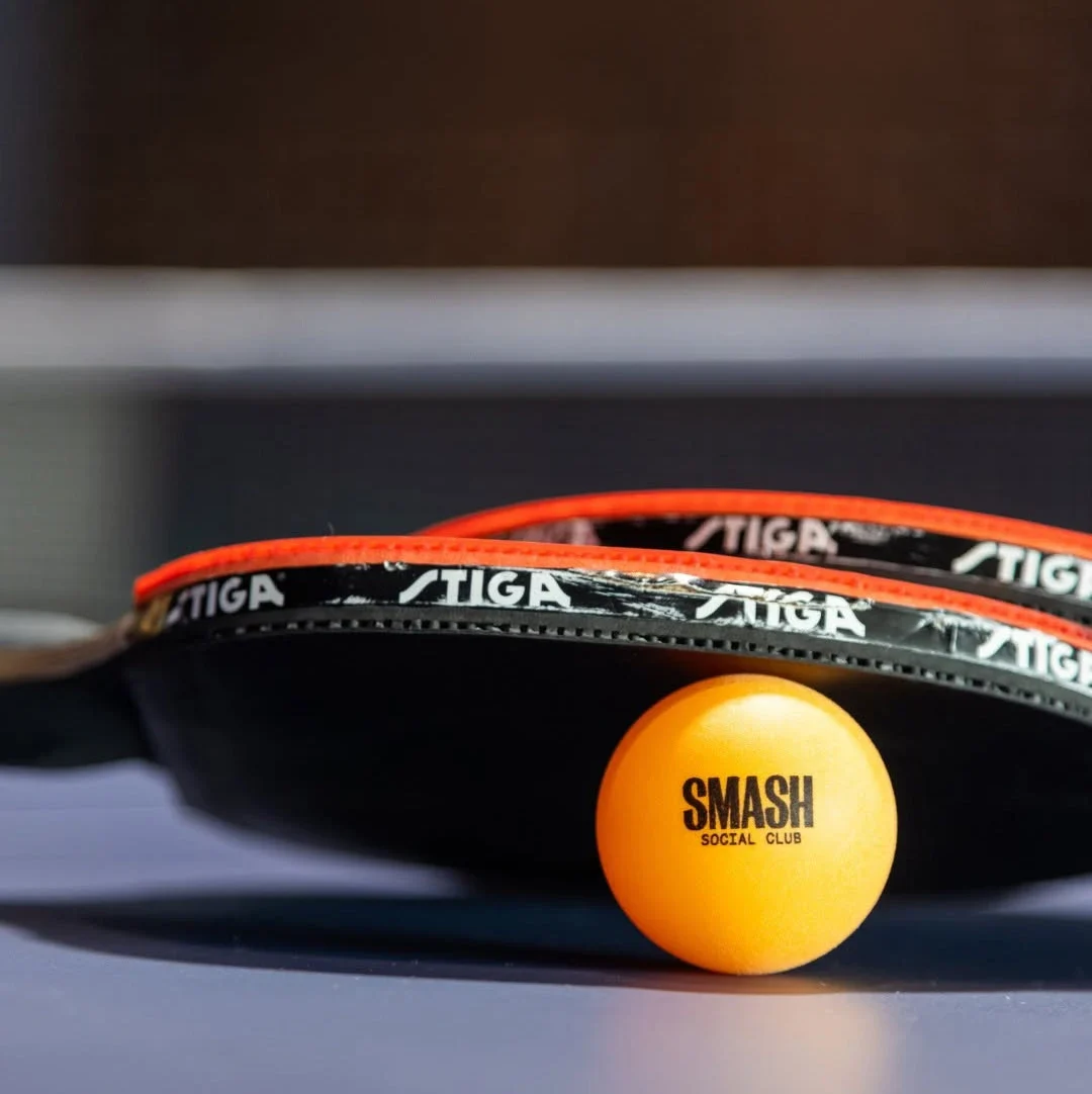 Two table tennis paddles rest on an orange ping pong ball with the SMASH Social Club logo printed on it