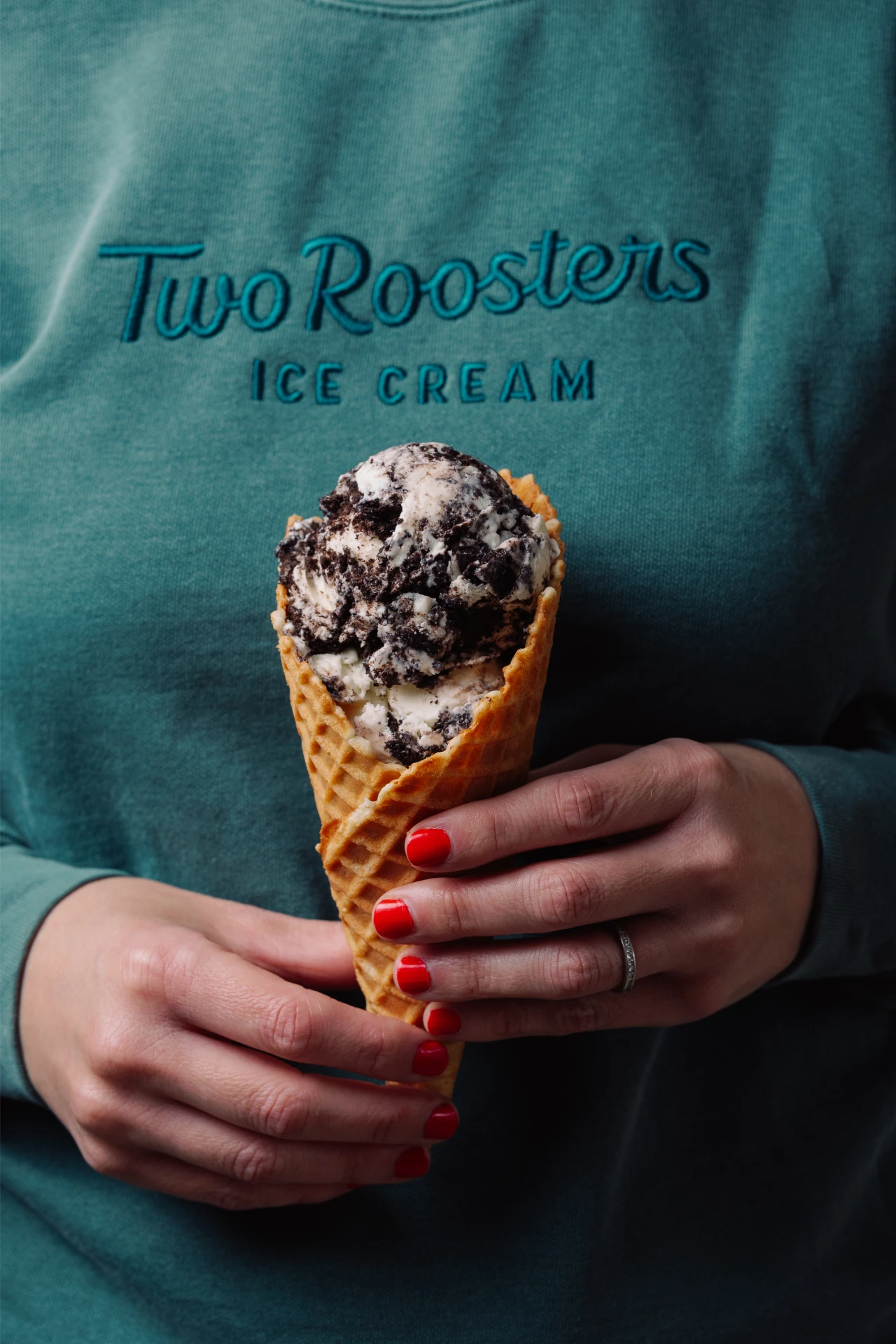 A woman wearing a Two Roosters logo embroidered sweatshirt and holding a waffle cone with ice cream