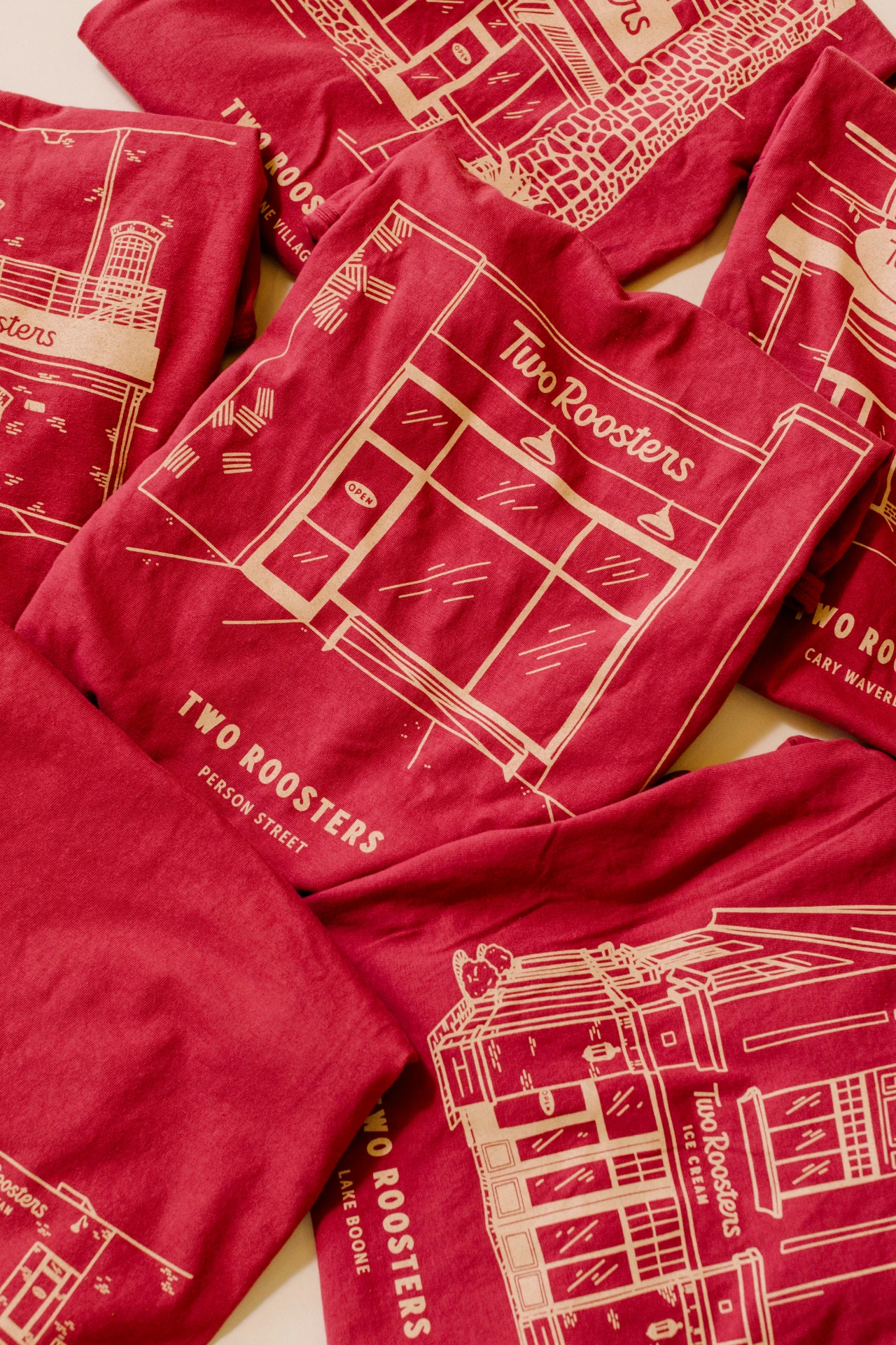 A pile of red t-shirts, each with a different Two Roosters storefront illustration