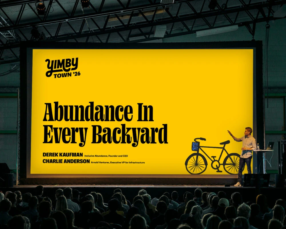 A presenter is on stage in front a large screen with a yellow background and the title Abundance in Every Backyard with the YimbyTown logo in the corner